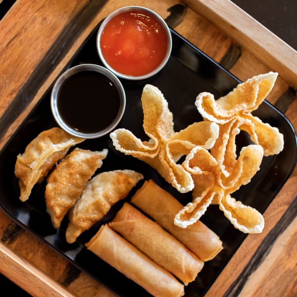 A black plate of Hibachi Boy appetizers, including gyoza, crab rangoons, and spring rolls with dipping sauces.